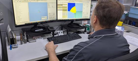 Engineer in a gray polo shirt working on dual monitors displaying 3D design software in a manufacturing office.